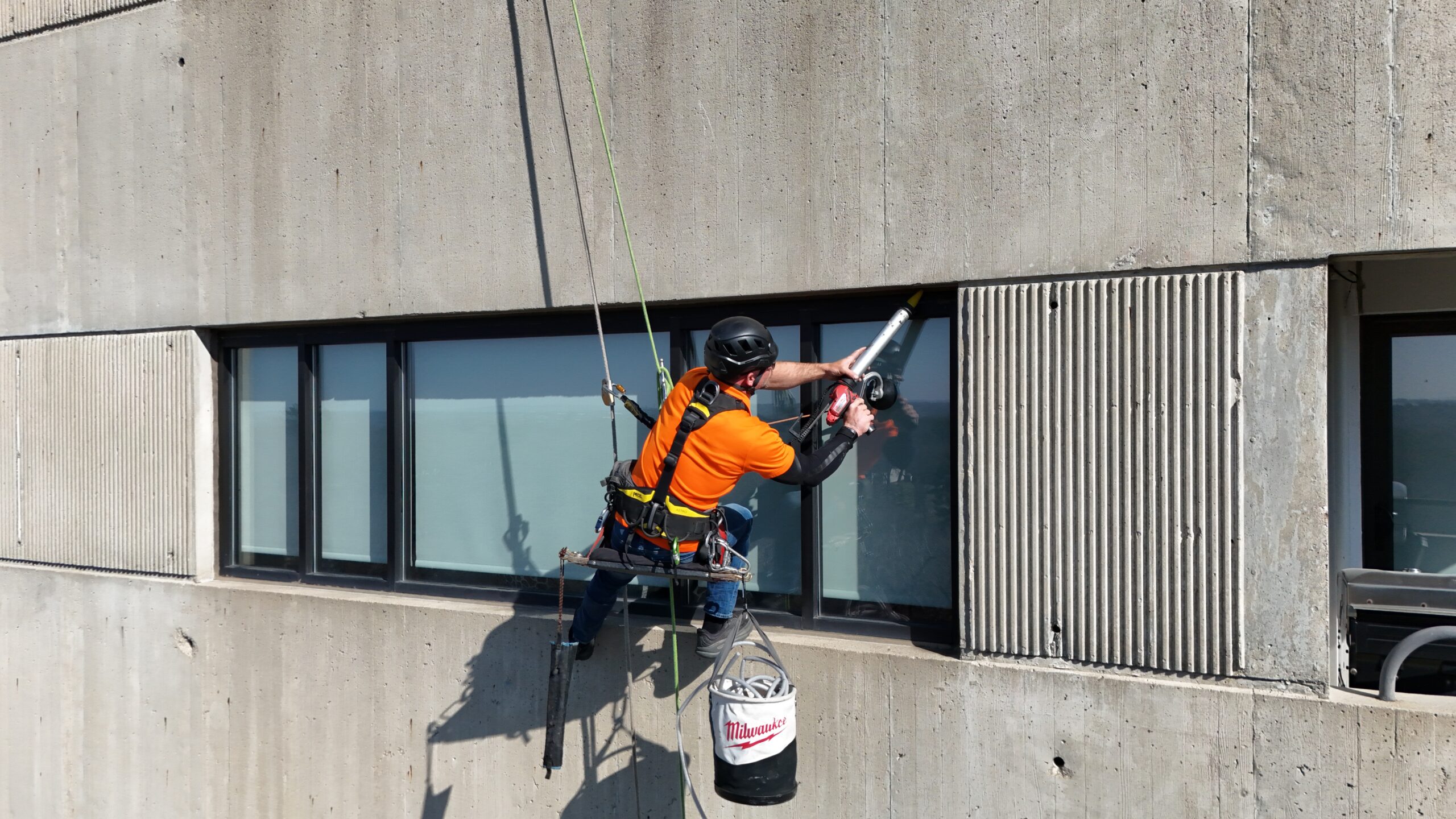 Worker in an orange shirt with black helmet performing rope-access caulking work on a commercial building in Des Moines, IA