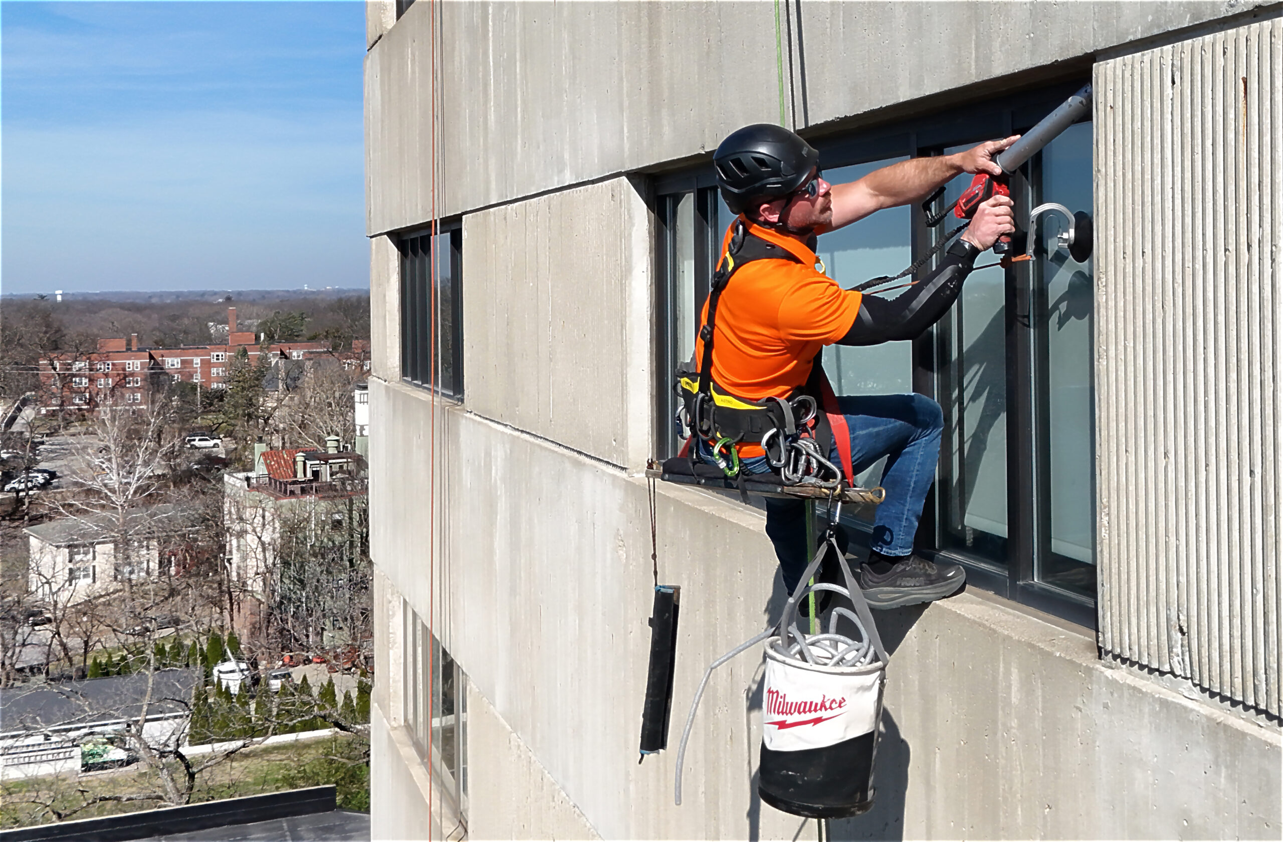 Closeup of a worker in an orange shirt with black helmet performing rope-access caulking work on a commercial building in Des Moines, IA