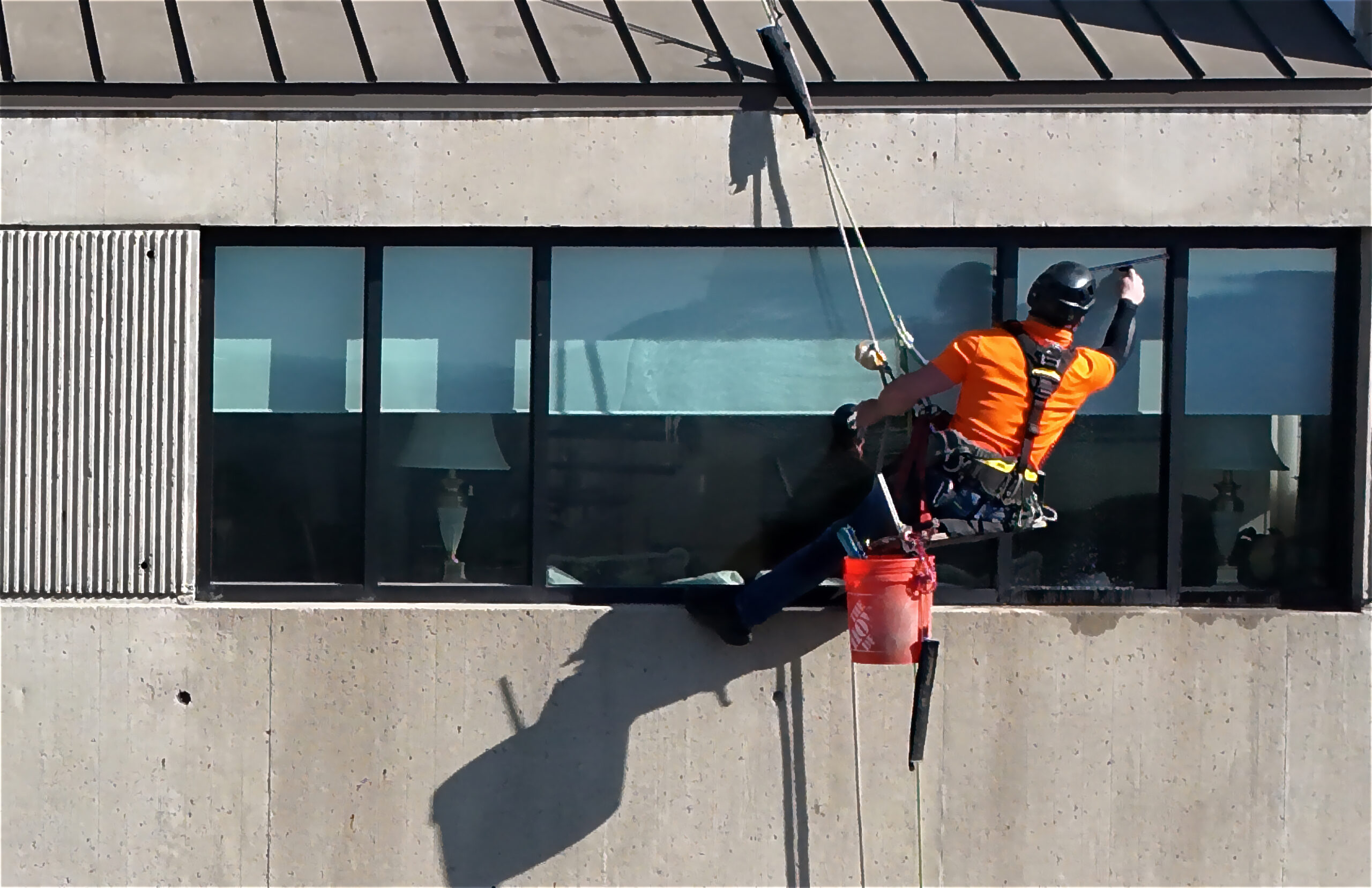 Closeup of a worker in an orange shirt with black helmet performing rope-access window cleaning on a commercial building in Des Moines, IA